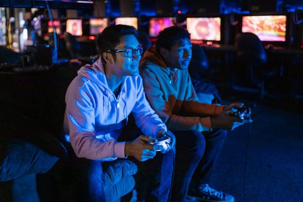 Two gamers in a Southeast Asian internet café locked in intense play, controllers in hand and illuminated by colourful gaming monitors