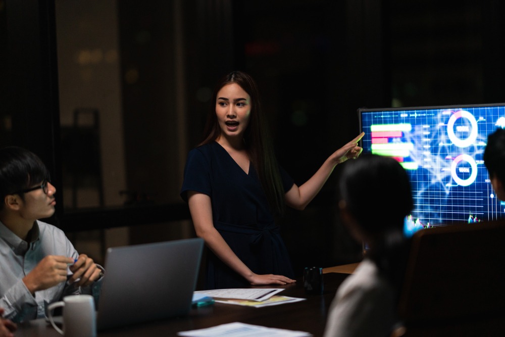 A female gaming marketing strategist presenting data and analytics on a screen to a team during a night meeting.