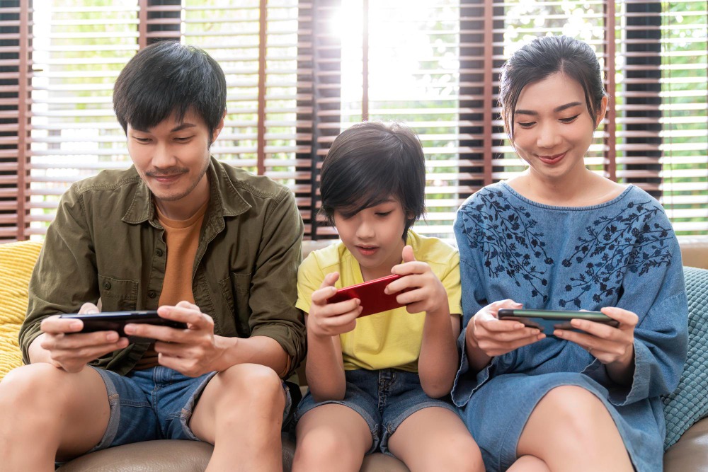 A Thai family sitting together on a couch playing mobile games on their smartphones, illustrating player engagement and retention.